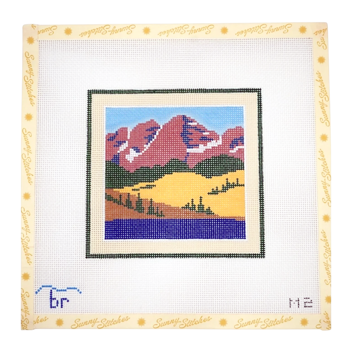 A framed needlepoint canvas featuring a Maroon Bells mountain landscape with blue sky, purple mountains, yellow hills, and a dark blue lake. The canvas is bordered with a cream-colored frame printed with the brand name 'Sunny Stitches' in gold.