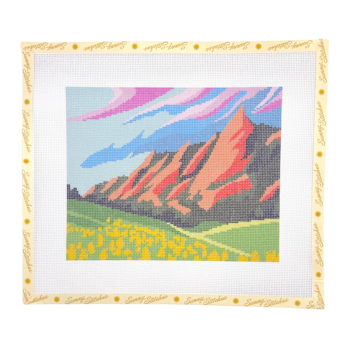 A needlepoint canvas featuring a colorful landscape of the Red Rocks with yellow wildflowers and a blue sky with pink clouds, surrounded by a white mesh border with a cream-colored stitched edge.