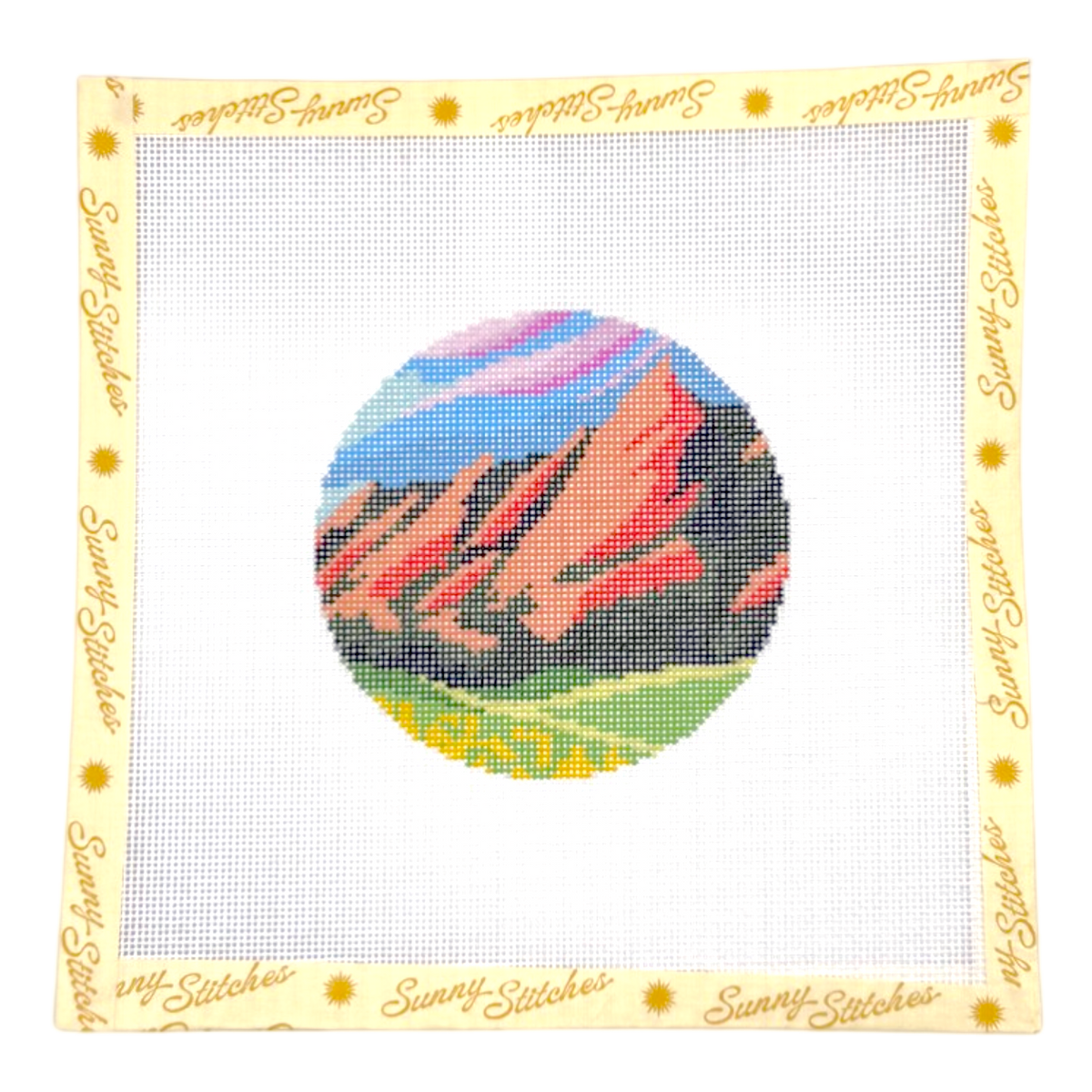 A square canvas needlepoint design by Sunny Stitches, featuring a circular landscape of red rock formations under a blue and pink sky with green fields. The white mesh is bordered by a cream-colored fabric edge printed with the brand name 'Sunny Stitches' in gold script.