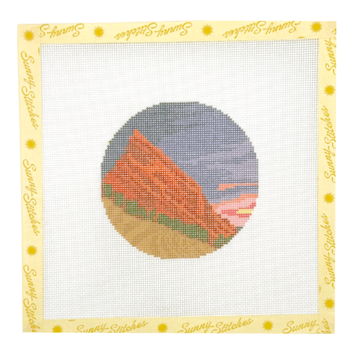A square needlepoint canvas with a white grid and a decorative border printed with the brand name 'Sunny Stitches' in yellow script. In the center is a circular painted design showing a red-orange mountain or rock formation against a blue and pink sky.